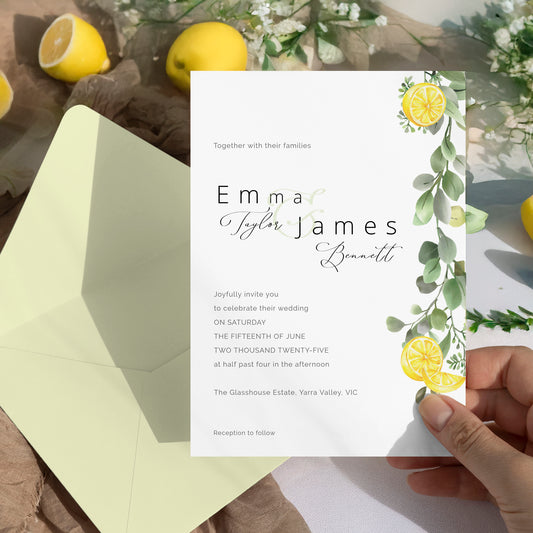 A wedding invitation card with watercolor lemon and eucalyptus greenery graphics, held by a hand over an open pale yellow envelope. The background is a rustic flat-lay with lemons and white flowers.