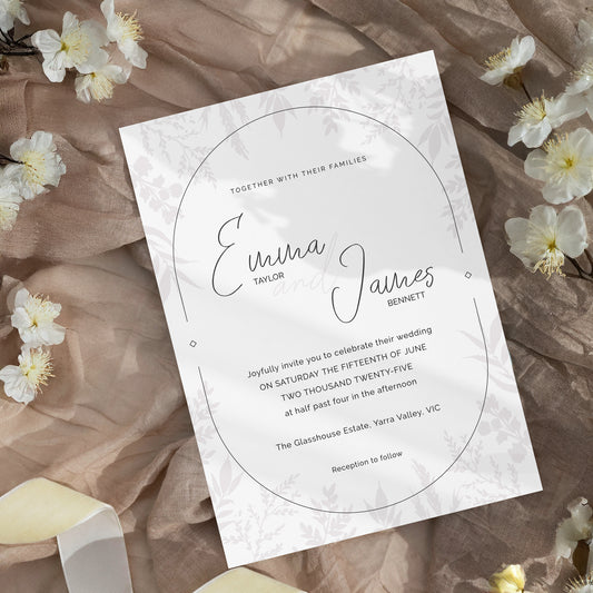 A wedding invitation card with elegant black script and serif text, featuring a circular frame design and a subtle floral pattern. The card is styled on a textured brown fabric background with scattered white flowers.