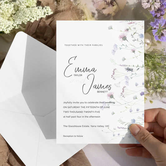 A hand holds a white wedding invitation card with elegant black script and floral illustrations, next to an open white envelope, styled with dried flowers and greenery.