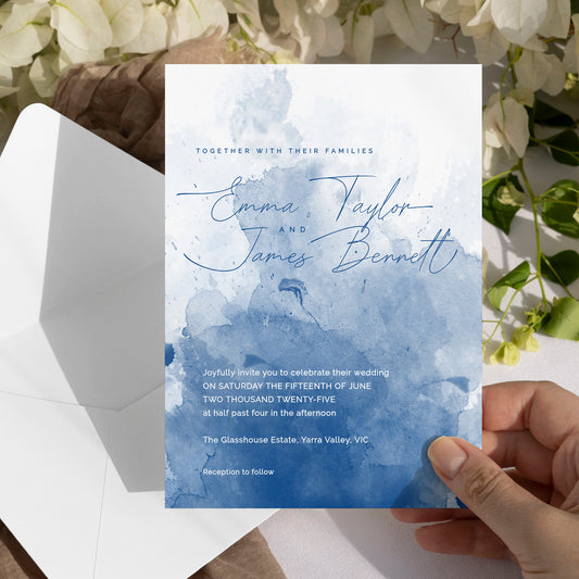 A hand holds a wedding invitation card featuring a blue watercolor landscape design, with an open white envelope and white flowers in the background.