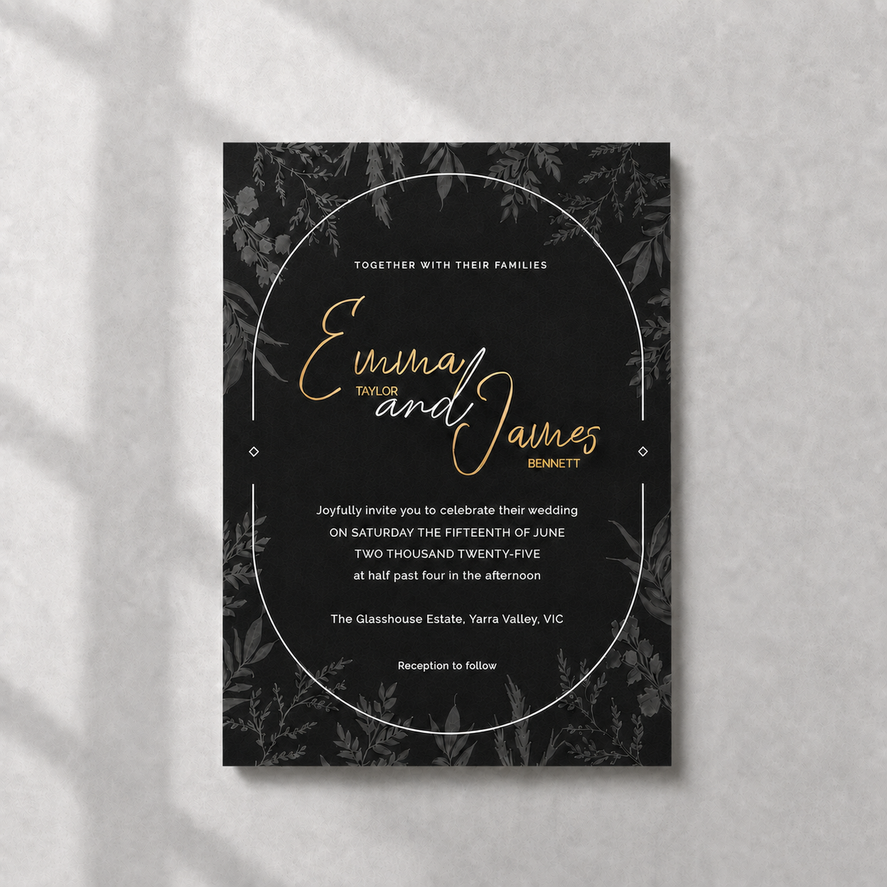 A rectangular wedding invitation card with a black background, subtle botanical border pattern, and elegant gold and white typography, photographed flat against a light textured wall with soft shadow overlays.