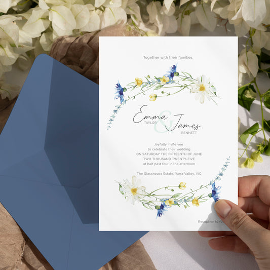 A wedding invitation card with floral watercolor designs is held by a hand, placed over an open blue envelope on a rustic surface with white flowers in the background.