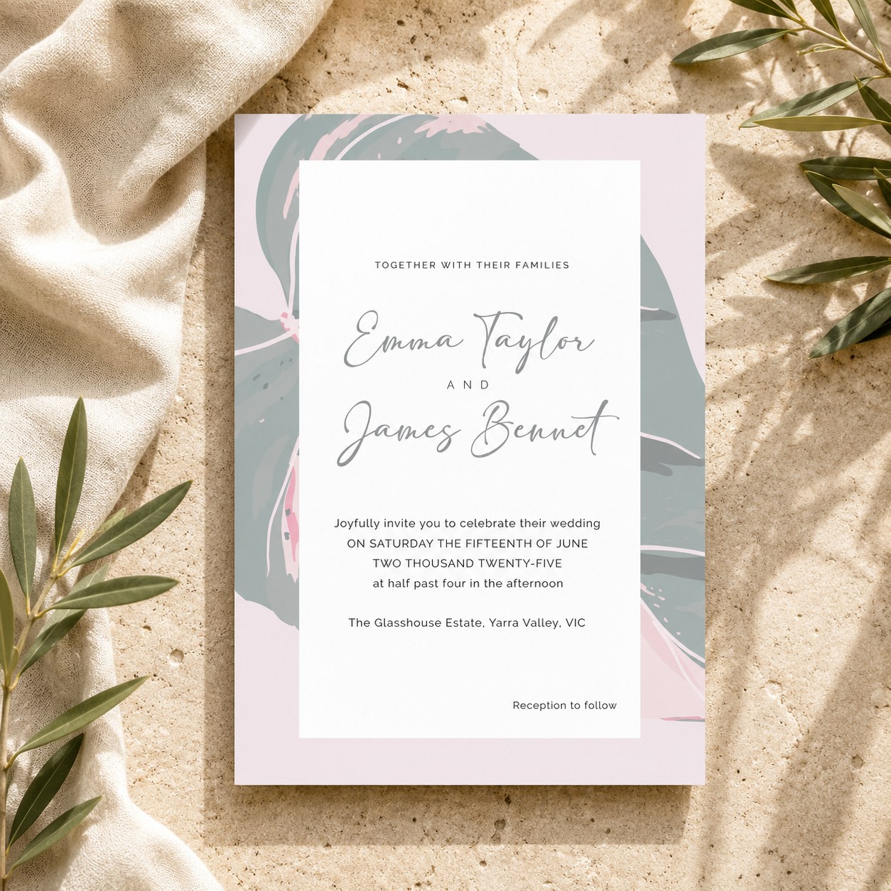 A wedding invitation card with a white center panel and abstract pastel artwork in pink, teal, and gray, styled on a sandy surface with draped beige fabric and olive branches.