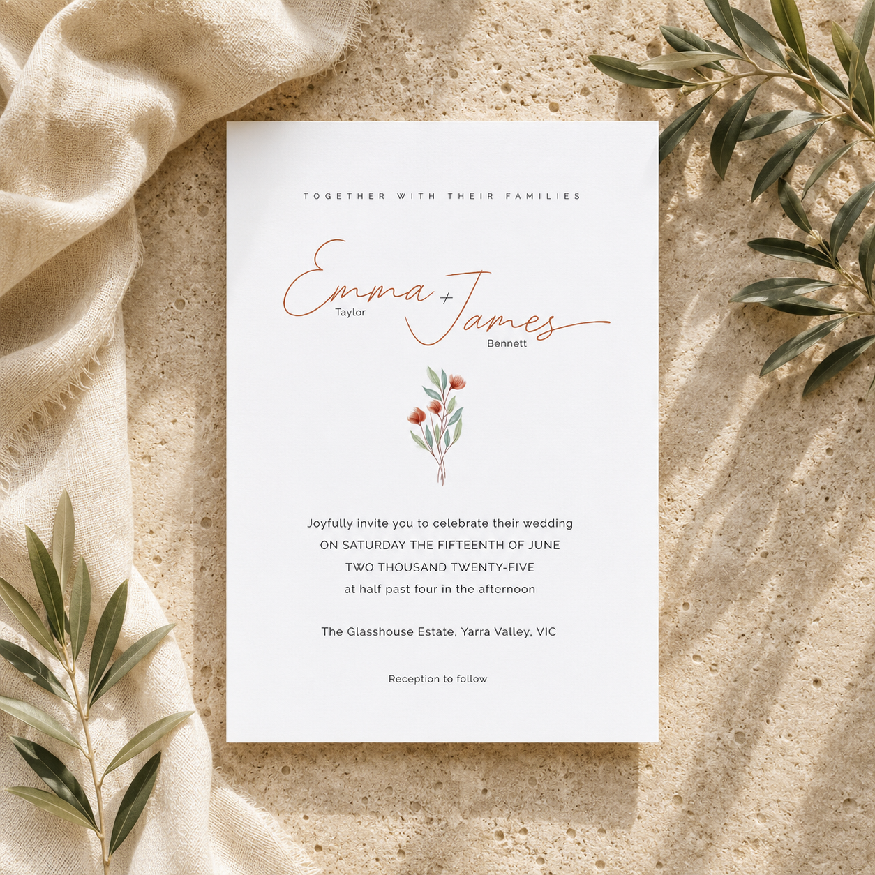 A wedding invitation card laid flat on a sandy surface with draped beige fabric and green leaves around it. The card is white with elegant copper script reading 'Emma + James' and smaller black text below.