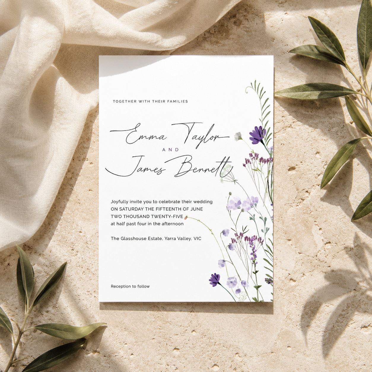 A rectangular wedding invitation card with elegant black script and floral illustrations in purple and green, styled flat-lay on a textured beige stone surface with draped cream fabric and olive leaves.