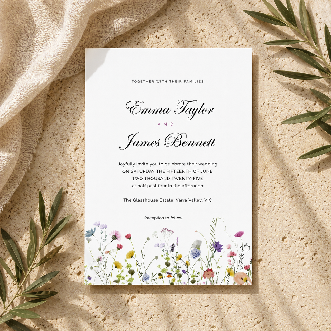 A wedding invitation card with elegant black script and floral watercolor illustrations at the bottom, styled flat-lay on a textured beige surface with draped cream fabric and green leaves.