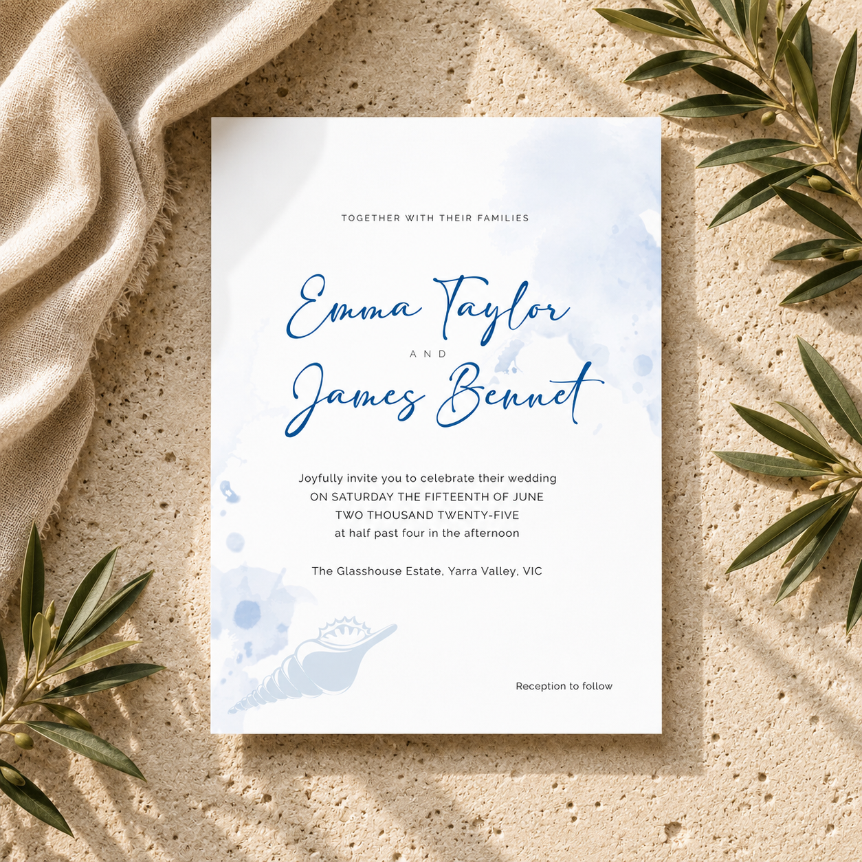 A wedding invitation card with elegant blue script and watercolor details, styled in a flat-lay on a sandy surface with draped beige fabric and green leaves.