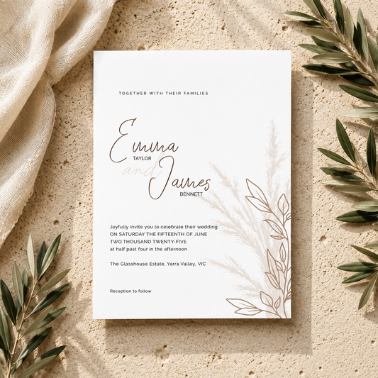 A wedding invitation card with elegant black script and serif text, styled in a flat-lay on a textured beige surface with draped cream fabric and olive branches.