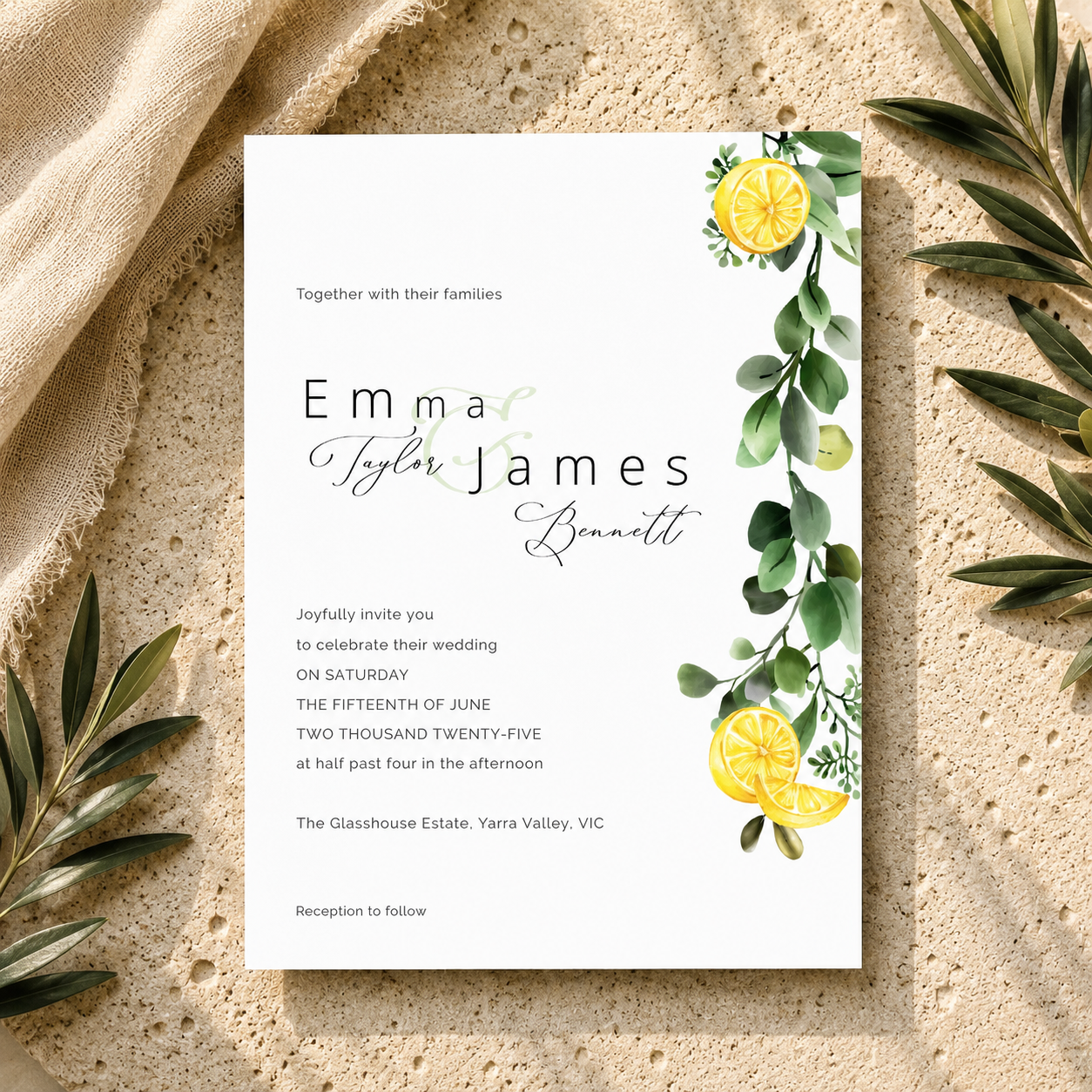 A wedding invitation card with elegant black and cursive text, personalized names, and watercolor lemon and eucalyptus illustrations, styled flat-lay on a textured beige surface with linen and olive leaves.