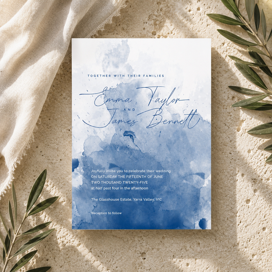 A wedding invitation card with elegant blue watercolor designs and script text reading 'Emma Taylor and James Bennett,' styled flat-lay on a textured beige surface with draped fabric and olive branches.