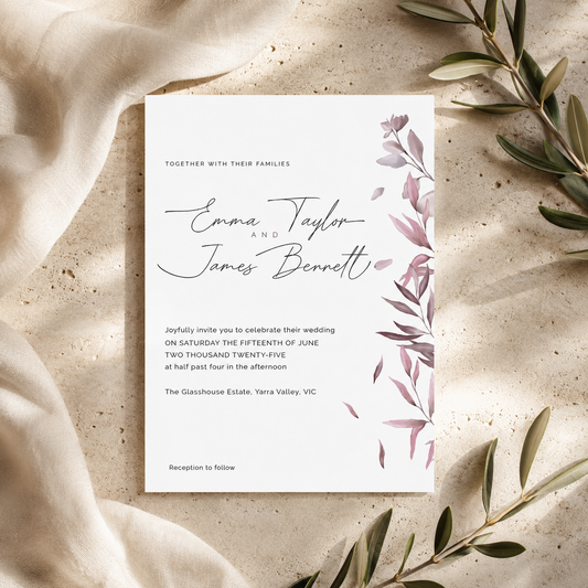 A wedding invitation card with elegant black script and gray text, decorated with a dusty pink botanical leaf border on the right side. The card is styled in a flat-lay arrangement on a textured beige surface with draped linen and olive branches.