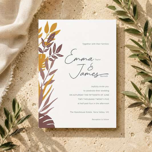 A square wedding invitation card with a white background, black script and serif text, and a botanical leaf illustration in mustard yellow, taupe, and brown along the left side. The card is photographed from above on a textured beige surface with draped cream fabric and green leaves around it.