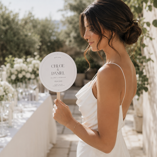 A woman in a white sleeveless dress stands outdoors holding a round printed sign on a stick. The sign reads 'CHLOE & DANIEL' with wedding event details.