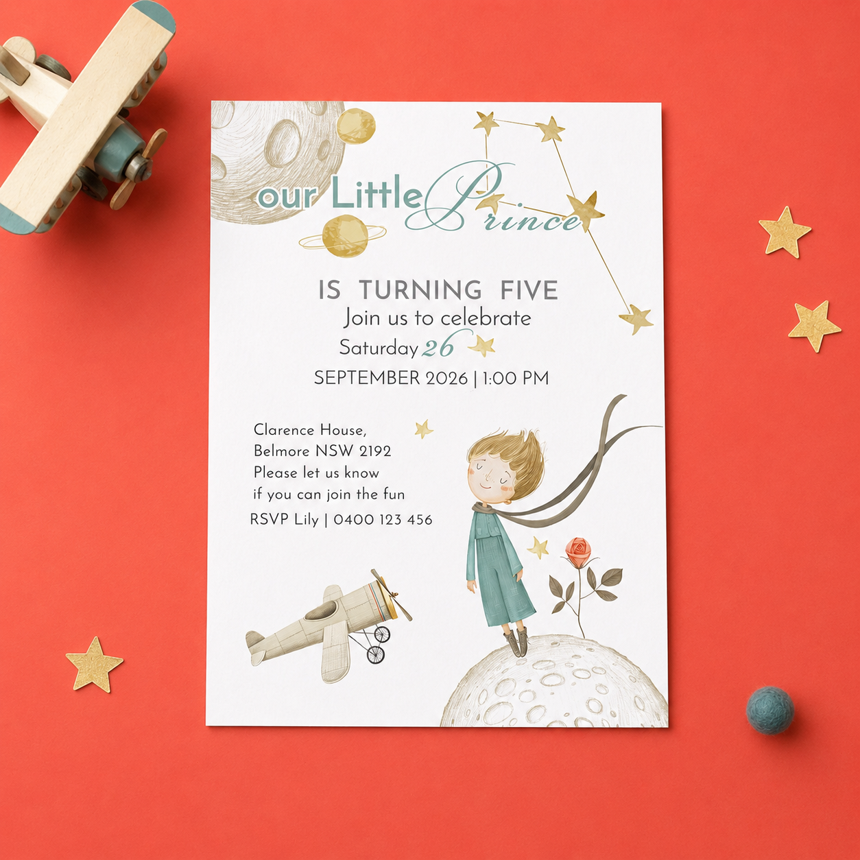 A children's birthday invitation card styled in a flat lay on a coral-red background, surrounded by toy airplane pieces, gold star cutouts, and a small blue ball. The card reads 'our Little Prince IS TURNING FIVE' with space-themed illustrations including a boy, moon, planets, and stars.