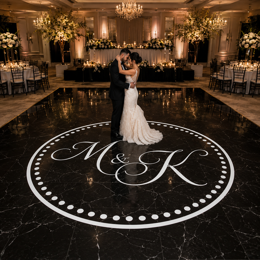 A bride in a white wedding gown and a groom in a black suit kiss on a polished black marble dance floor featuring a large white circular monogram design. The background shows a grand, elegantly decorated wedding reception hall with chandeliers, floral arrangements, and dining tables.