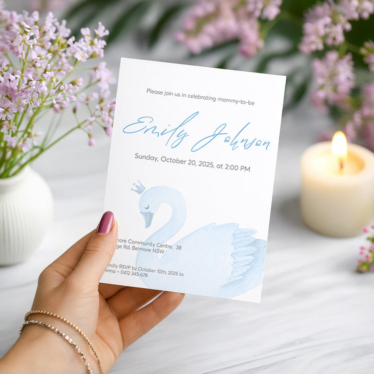 Hand holding a baby shower invitation with a swan design, surrounded by flowers and a candle.