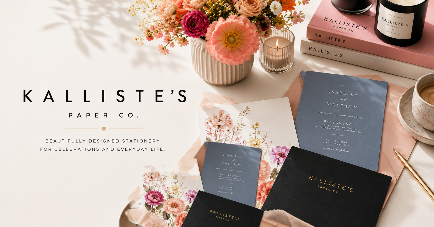 A styled flat-lay of Kalliste's Paper Co. stationery items on a light neutral surface, including blue and black invitation cards, a floral card, a pink book, candles, flowers, and a cup of coffee.