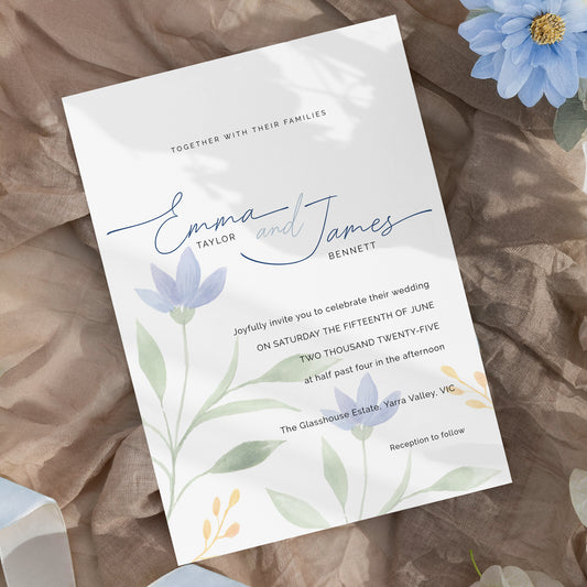 A wedding invitation card featuring elegant script names 'Emma and James Bennett' with floral watercolor illustrations in soft blue, green, and yellow, styled on a textured brown fabric background with decorative blue flowers.