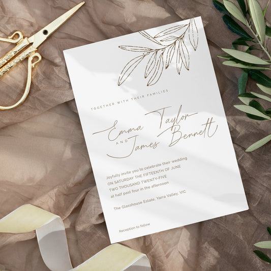 A wedding invitation card with elegant gold foil script and leaf illustrations, styled on a brown textured fabric with gold scissors, ribbon, and green leaves.