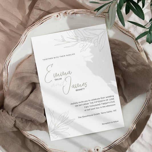 A wedding invitation card with elegant botanical line art, styled on a rustic white wooden tray with neutral fabric and green leaves.