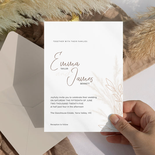A white wedding invitation card held by a hand, featuring elegant black script names and delicate botanical line art, styled with a matching envelope and dried pampas grass.