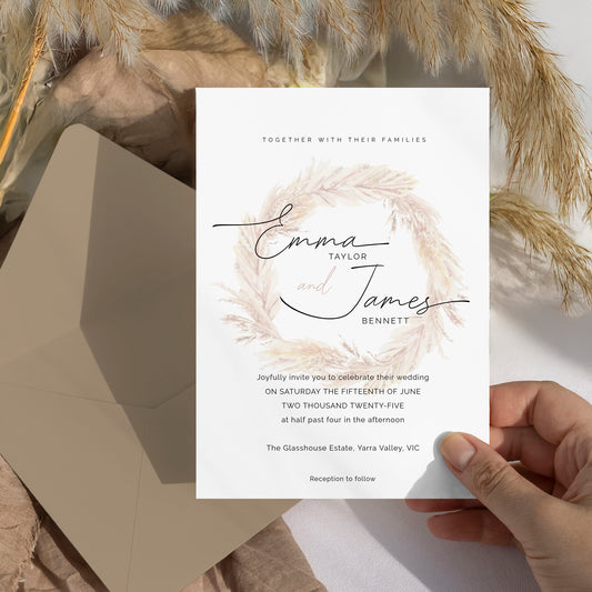 A wedding invitation card held by a hand, featuring a watercolor feather wreath design with the names 'Emma and James Bennett' in black script. The card is styled with a kraft envelope and dried pampas grass in a neutral, rustic setting.