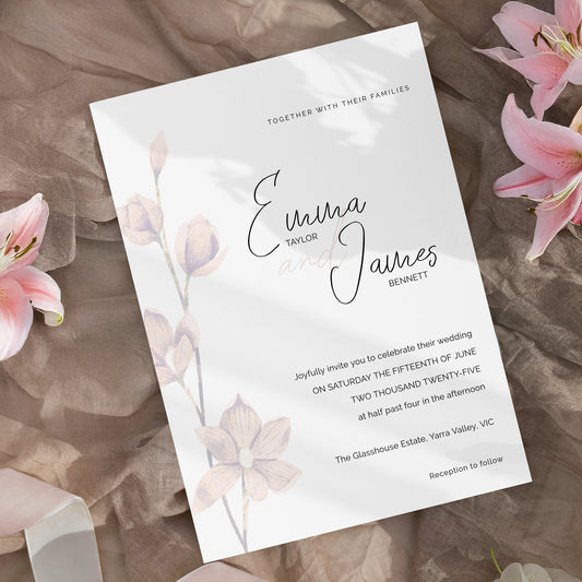 A wedding invitation card with elegant black script and gray text, decorated with soft pink watercolor flowers, displayed on a textured brown fabric background with pink lilies.