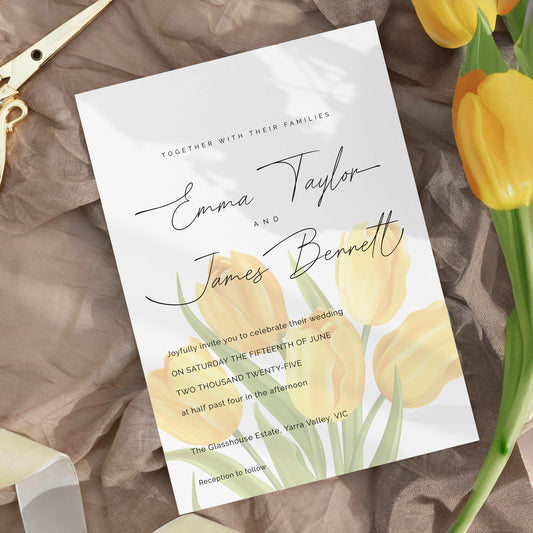 A wedding invitation card styled in a flat-lay with yellow tulips and gold scissors on a brown fabric background. The card features elegant black script text reading 'Emma Taylor and James Bennett' with yellow tulip illustrations at the bottom.