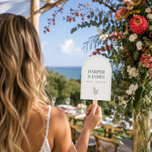 A woman at a wedding holds a personalized table sign that reads 'HARPER & JAMES' with a scenic outdoor view of the ocean and floral arch in the background.