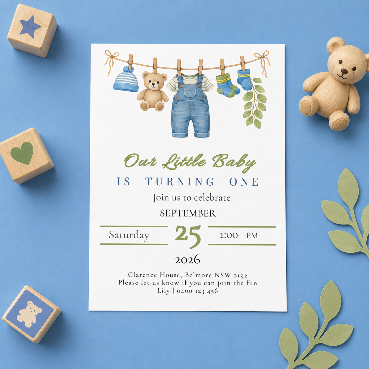 A baby boy's first birthday invitation card styled in a flat lay on a blue background with wooden toy blocks, a teddy bear, and paper leaves. The card features a watercolor illustration of baby clothes hanging on a clothesline.
