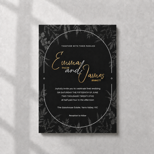 A rectangular wedding invitation card with a black background, subtle botanical border pattern, and elegant gold and white typography, photographed flat against a light textured wall with soft shadow overlays.