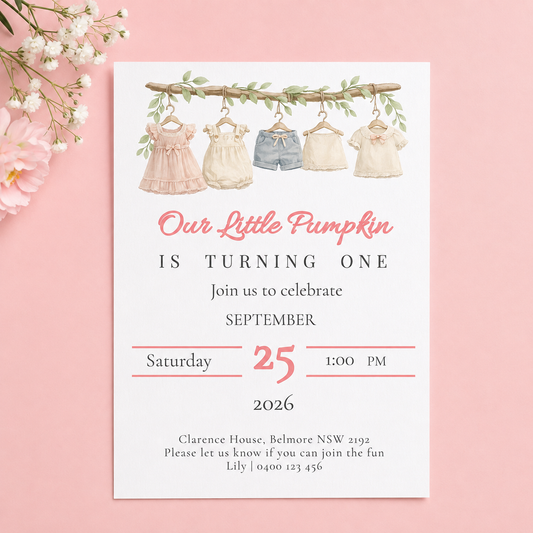 A baby first birthday invitation card styled on a pink background with white flowers. The card features watercolor illustrations of four baby dresses hanging on a clothesline, with the text 'Our Little Pumpkin IS TURNING ONE' in pink and black lettering.