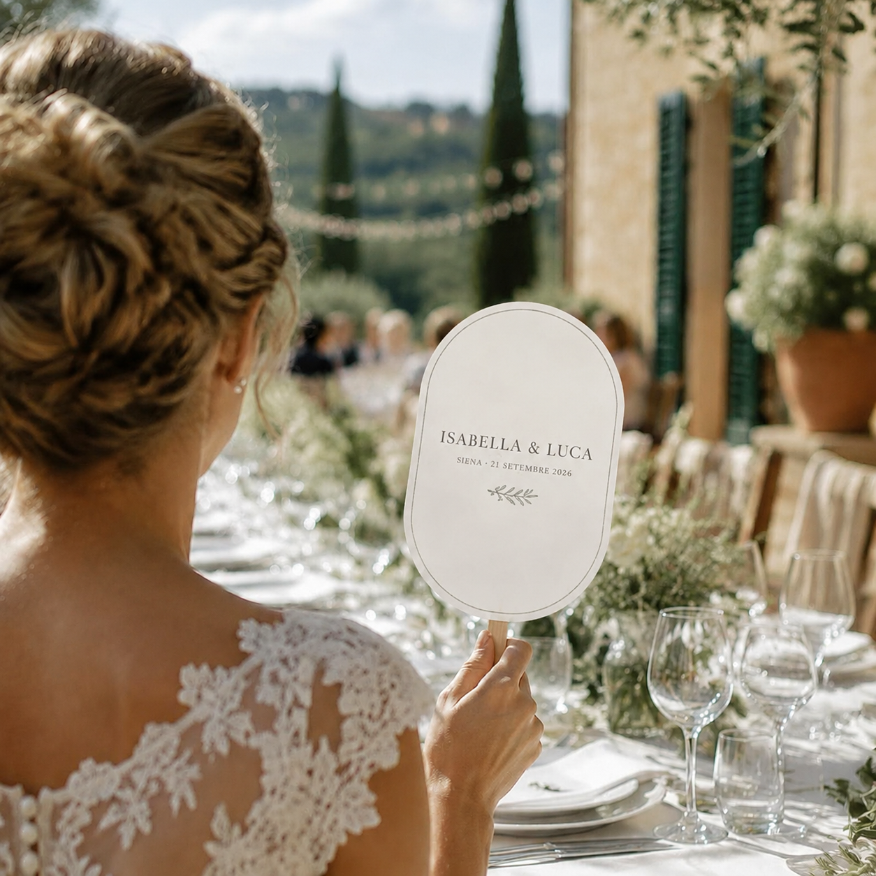 A bride in a white lace wedding dress holds an oval white guest sign with personalized wedding text, photographed from behind in an elegant outdoor wedding setting with tables, wine glasses, and greenery.