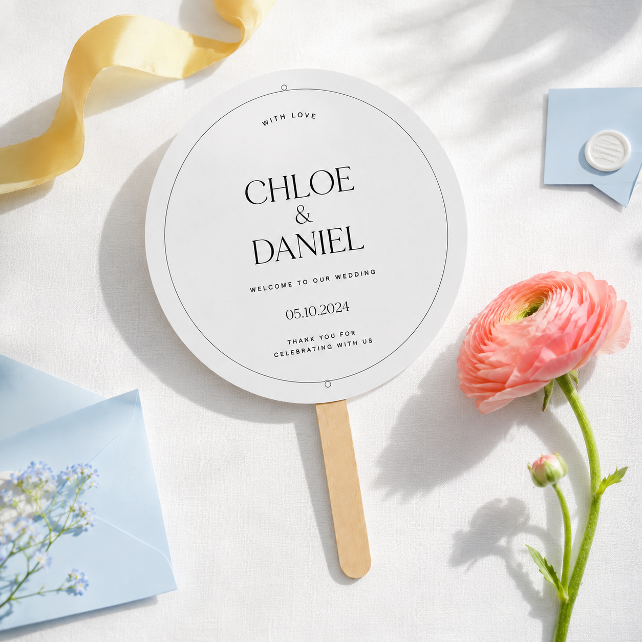 A personalized wedding fan sign with a wooden handle, printed with 'CHLOE & DANIEL' and wedding details, styled on a white surface with a pink ranunculus flower, a yellow ribbon, and light blue stationery.