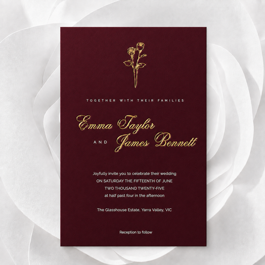 A rectangular wedding invitation card with a deep burgundy background and elegant gold typography, centered on a white floral backdrop. The card includes a small gold rose illustration at the top and names Emma Taylor and James Bennett.