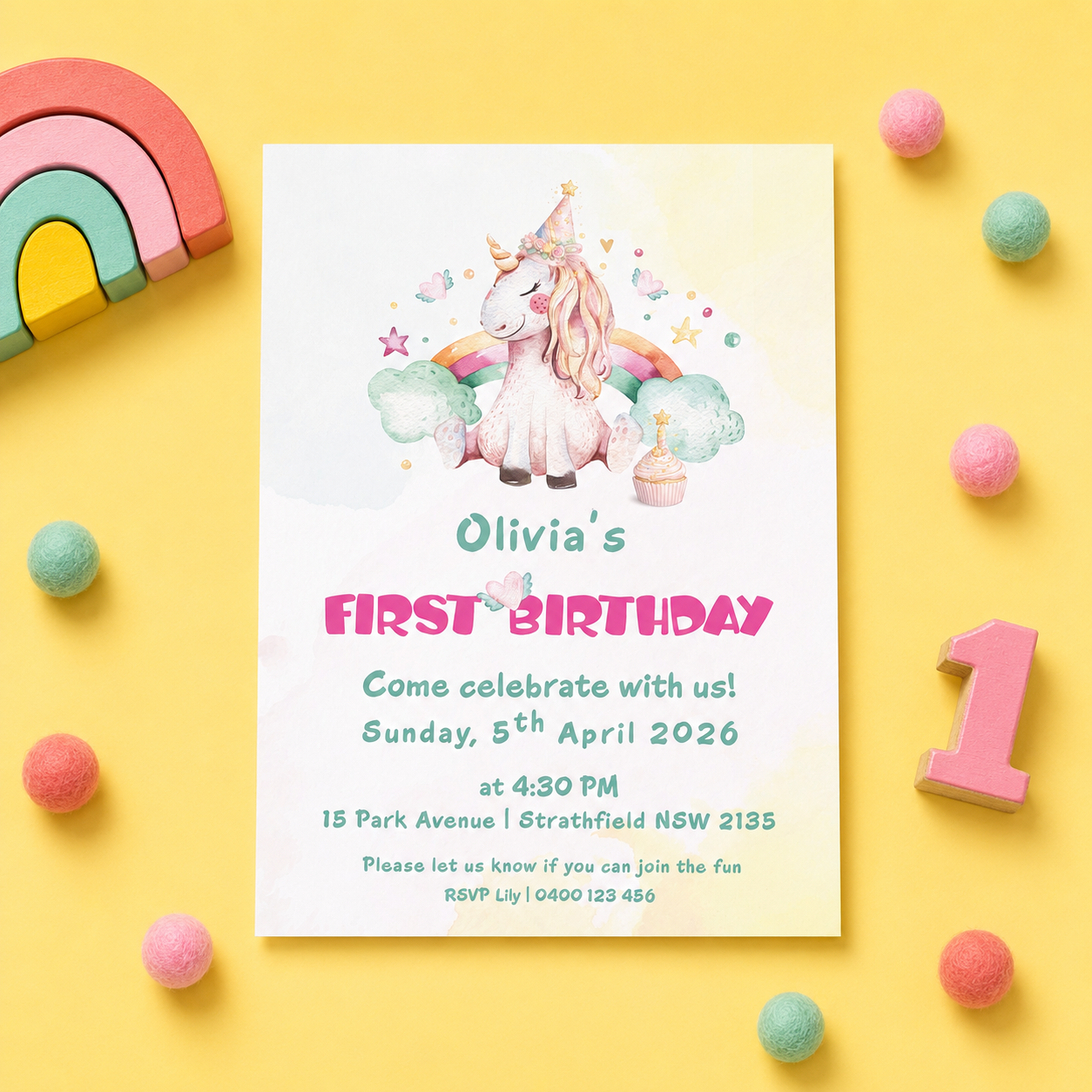 A pastel unicorn first birthday invitation card styled in a flat lay on a yellow background with colorful wooden rainbow blocks, pink pom-poms, a pink number 1 block, and a wooden spoon.