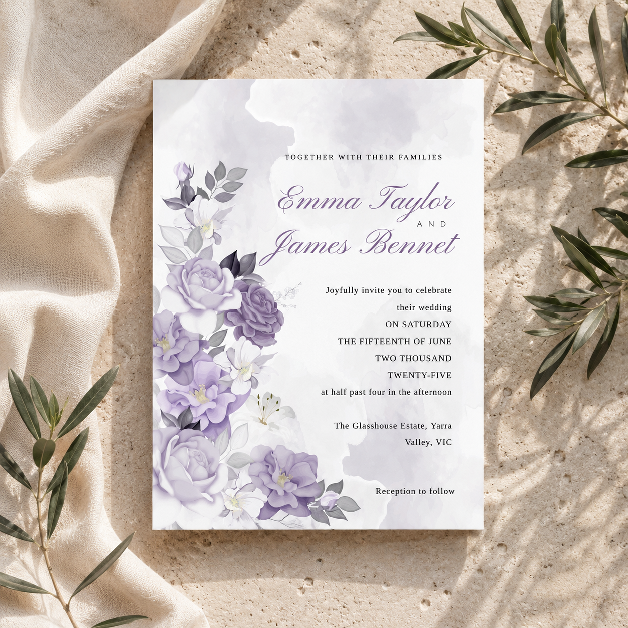 A floral wedding invitation card with lavender and purple watercolor flowers, placed on a textured beige fabric surface with olive branches around it.