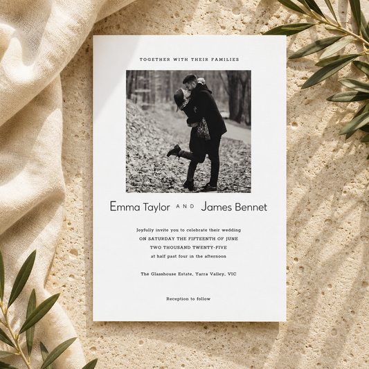 A wedding invitation card laid flat on a textured beige surface with cream fabric and green leaves around it. The card has a black-and-white family photo at the top and printed text for Emma Taylor and James Bennet.