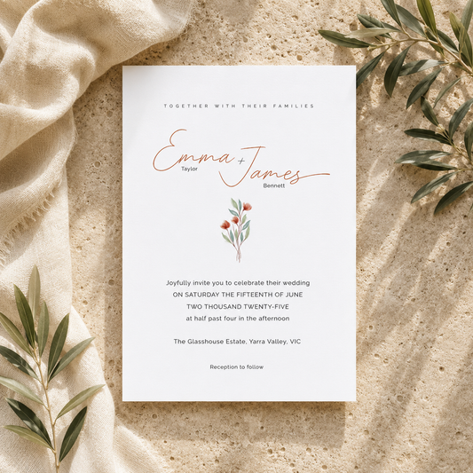 A wedding invitation card laid flat on a sandy surface with draped beige fabric and green leaves around it. The card is white with elegant copper script reading 'Emma + James' and smaller black text below.