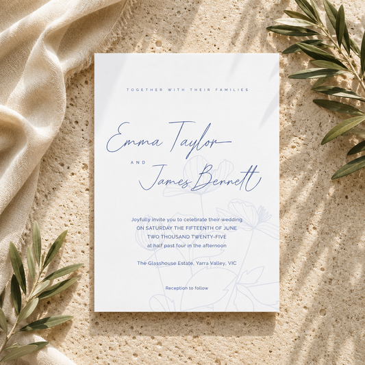 A wedding invitation card with elegant blue script and floral line art, styled flat-lay on a textured beige stone surface with draped cream fabric and olive branches.