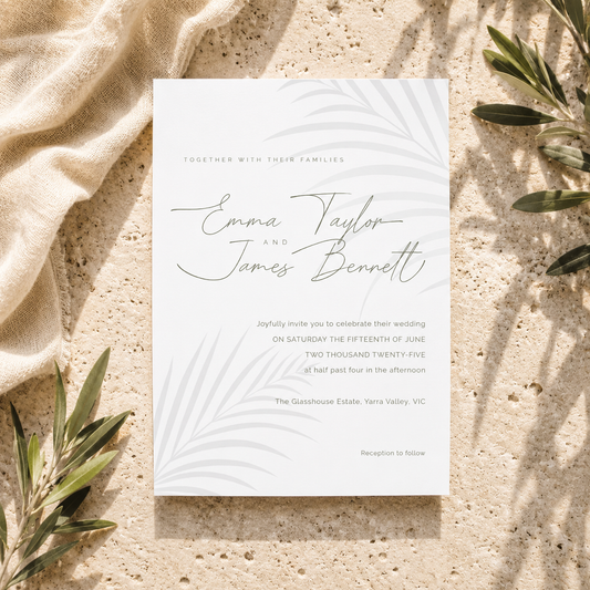 A wedding invitation card with elegant gray script and leafy line art, styled flat-lay on a textured beige stone surface with draped linen and olive branches.