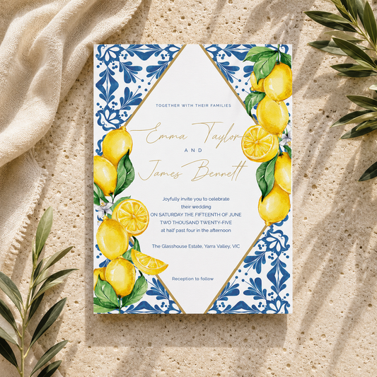 A wedding invitation card with a white center, blue floral border, and yellow lemon illustrations, photographed flat on a sandy surface with draped fabric and green leaves around it.