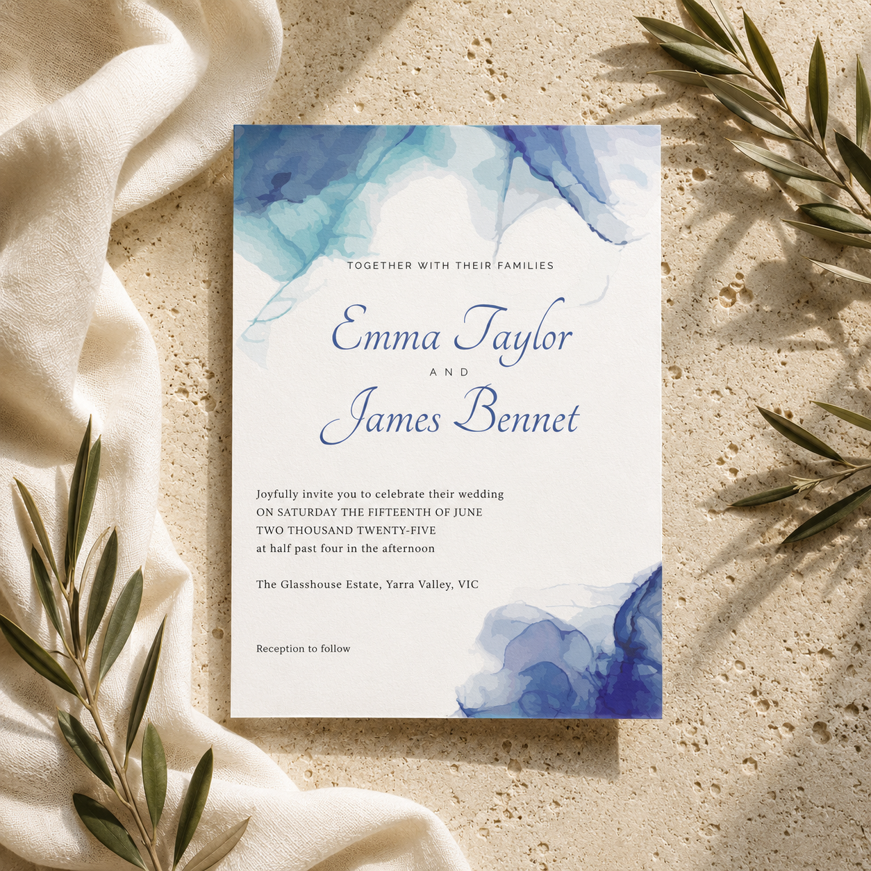 A square wedding invitation card with blue watercolor floral designs in the corners, laid flat on a textured beige stone surface with draped cream fabric and olive branches around it. The card reads 'Together with their families Emma Taylor and James Bennet' and includes wedding event details.