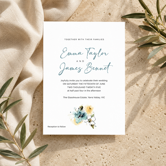 A wedding invitation card with elegant blue script text and a small floral illustration, styled on a beige textured surface with draped cream fabric and green leaves around it.