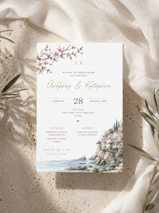 A square wedding invitation card with elegant typography, pink floral illustrations, and a coastal village scene at the bottom, styled on a textured beige surface with draped cream fabric and olive branches.