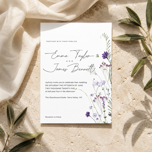 A rectangular wedding invitation card with elegant black script and floral illustrations in purple and green, styled flat-lay on a textured beige stone surface with draped cream fabric and olive leaves.