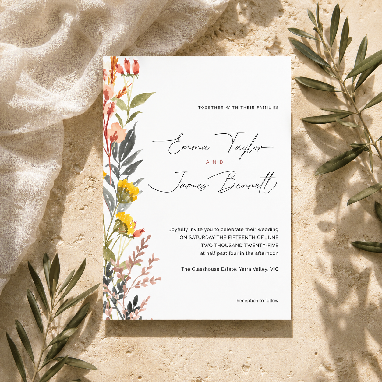 A rectangular wedding invitation card with floral watercolor illustrations in red, orange, yellow, and green, laid flat on a light beige textured surface with draped white fabric and olive branches around it.