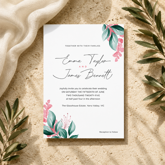 A square wedding invitation card with elegant black script and floral watercolor accents in teal and pink, styled flat-lay on a textured beige surface with draped cream fabric and green leaves around it.