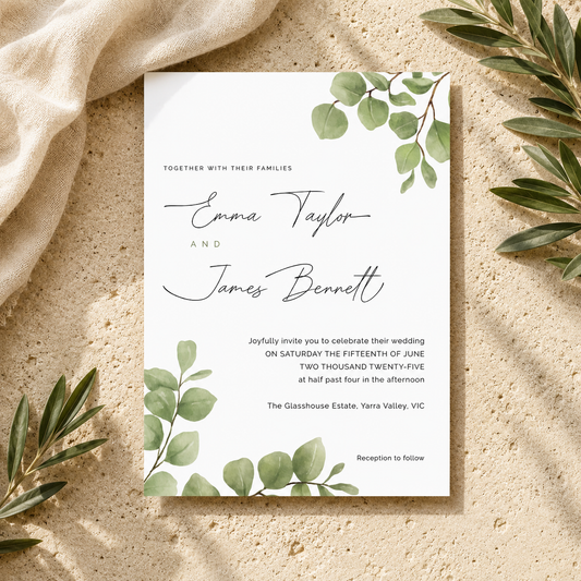 A rectangular wedding invitation card with elegant black script and serif text, decorated with green eucalyptus leaves at the top and bottom, styled on a textured beige surface with draped cream fabric and olive branches.