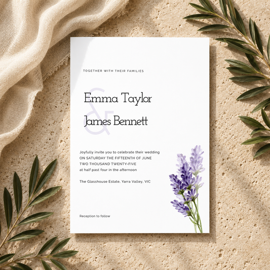 A square wedding invitation card with elegant black typography and a small purple floral illustration in the lower right, styled on a textured beige surface with draped cream fabric and green leaves around it.